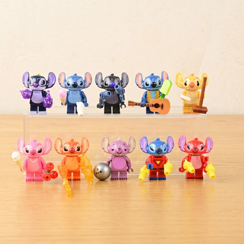 May include: A collection of colorful Stitch character figurines, each in a unique pose and holding different accessories. The figurines are arranged on a clear display shelf, with a light wooden surface in the background.