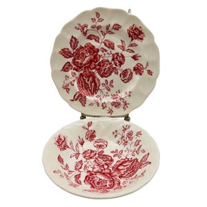 May include: Two cream-colored ceramic plates with scalloped edges, each featuring a detailed red floral pattern of roses and leaves. The plates are displayed vertically, one positioned above the other, showcasing the intricate botanical design.