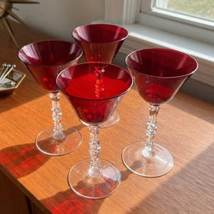 May include: A set of four cocktail glasses featuring deep red, conical bowls and clear, faceted stems with circular bases. The glass bowls are wide at the top and taper down to the stem, creating a distinct silhouette for serving beverages.