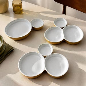 May include: Three ceramic serving dishes with a white interior and a mustard yellow exterior. One dish is oval with a small circular side compartment, while the other two are larger, featuring two circular main sections and one smaller circular compartment.