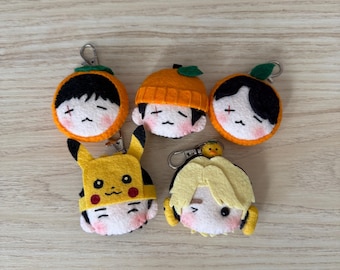 May include: Five felt plush keychains featuring stylized character faces. Three are topped with orange fruit designs, one wears a yellow Pikachu-inspired hood, and one features blonde hair with a small yellow bird perched on top.