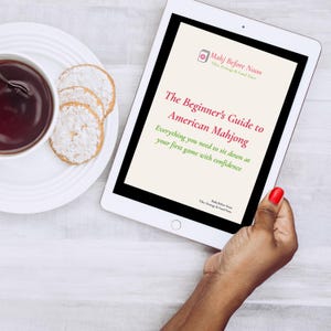 May include: A tablet displays "The Beginner's Guide to American Mahjong." The screen is held by a hand with red nail polish. A cup of tea and cookies are on a white plate to the left. The tablet's title is in a red font.