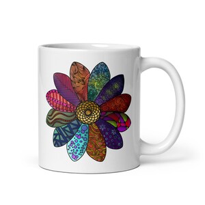May include: A white ceramic mug featuring a central flower graphic with twelve distinct petals. Each petal displays a unique, intricate pattern in vibrant colors including red, blue, green, purple, and orange, surrounding a golden yellow center.