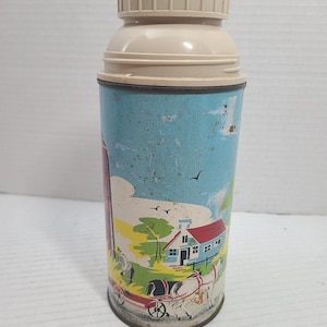 May include: A vintage metal thermos with a beige plastic cap and a red top button. The cylindrical body features a colourful illustration of a rural farm scene, including a house with a red roof, a horse-drawn carriage, and a red silo against a blue sky.