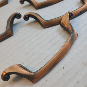 Puede incluir: Una colección de tiradores de cajón de metal en tono cobre con un diseño curvo y alargado con extremos en espiral. Cada tirador tiene un acabado de pátina oscura y desgastada, mostrando una textura metálica distintiva.