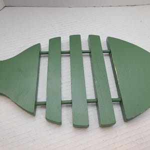 Puede incluir: Un salvamanteles de madera en color verde salvia con forma de pez, construido con cinco listones verticales unidos por dos varillas horizontales. La superficie muestra un ligero desgaste de pintura en los bordes y una forma definida de cola y cabeza.