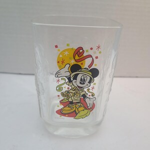 May include: A clear glass tumbler featuring a colorful graphic of Mickey Mouse dressed in a tan safari outfit with binoculars. The design includes a yellow sun, red ribbons, and green confetti accents. The word Disney is printed at the bottom.