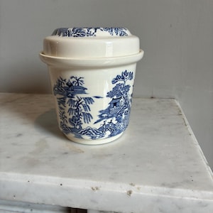 May include: A cream-colored ceramic lidded jar featuring a blue transferware pattern depicting a landscape with trees, a bridge, and buildings. The cylindrical container has a matching lid with a stepped design and a flared rim.