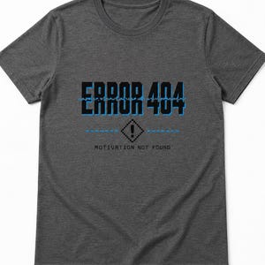 Op de afbeelding: Een antracietgrijs T-shirt met korte mouwen en de tekst ERROR 404 in het zwart met een blauwe omlijning. Onder de tekst staat een ruitvormig icoon met een uitroepteken en de tekst MOTIVATION NOT FOUND in een zwart pixelachtig lettertype.