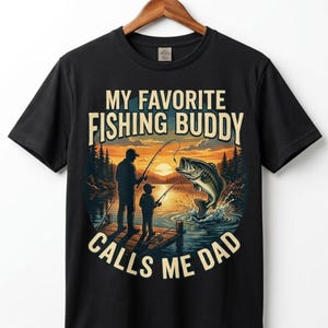 May include: A black short-sleeved t-shirt featuring a graphic print of an adult and a child fishing on a wooden dock at sunset. The text reads MY FAVORITE FISHING BUDDY CALLS ME DAD. The illustration includes a large fish jumping out of the water.