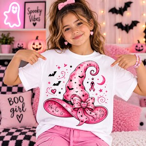 以下が含まれることがあります： 大きなピンク色の魔女の帽子のグラフィックがプリントされた白いTシャツ。デザインには、黒いコウモリ、ピンクの星、三日月、ピンクのリボン、中央にハートがあしらわれたリボンが含まれています。帽子は先がカールした尖った形状で、つばが広くなっています。