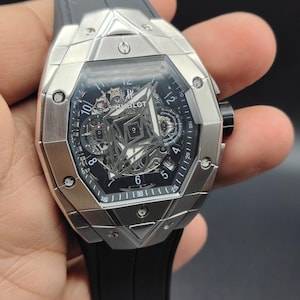 Könnte beinhalten: Eine Hublot-Armbanduhr mit einem silberfarbenen, kantigen Metallgehäuse mit sichtbaren Schrauben und einem skelettierten Zifferblatt. Die Uhr hat ein schwarzes Kautschukarmband und zeigt das Hublot-Logo sowie Ziffern auf dem Zifferblatt.
