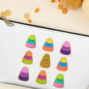 May include: A white zippered pouch featuring a repeating pattern of nine colorful, smiling candy corn-shaped icons. The text UPGRADE YOUR BEAUTY BAG appears at the top, and the logo for SUN, CANVAS, & HUE DESIGNS is at the bottom of the image.