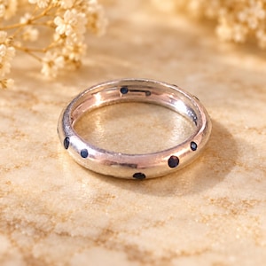 May include: A polished silver band ring featuring a series of small, round, dark blue gemstones set at regular intervals around the outer circumference of the metal band, creating a simple and consistent circular design.
