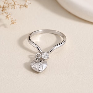 May include: A silver-toned ring featuring a V-shaped band with a single round clear stone set at the center. A heart-shaped charm hangs from the setting, decorated with fine etched lines and a vertical row of small clear stones along its side.