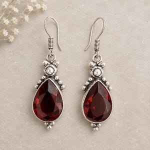 Puede incluir: Un par de pendientes colgantes de tono plateado con piedras de color rojo oscuro talladas en forma de pera. Los engastes de metal presentan pequeñas cuentas plateadas y detalles decorativos, suspendidos de ganchos simples de plata.