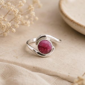Puede incluir: Un anillo de plata con una piedra preciosa redonda y pulida de color rosa intenso, engastada en un bisel de metal liso. La banda del anillo tiene un diseño cruzado con dos brazos de plata curvados que rodean la piedra central.