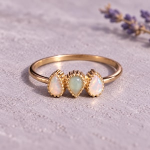 May include: A gold-toned ring featuring three pear-shaped stones arranged horizontally. The center stone is a pale green, while the two flanking stones are iridescent white opals. Each stone is set in a delicate, beaded gold-toned metal frame.