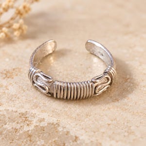 May include: A silver-toned open ring featuring a central section wrapped in thin wire coils, flanked by two decorative S-shaped wire loops on either side. The metal has a textured, slightly weathered finish throughout the entire band structure.