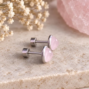 Puede incluir: Un par de pendientes de botón con piedras de cuarzo rosa triangulares y facetadas engastadas en metal plateado pulido. Cada pendiente tiene un poste recto y un cierre de rosca circular y plano, con un diseño limpio y sencillo.
