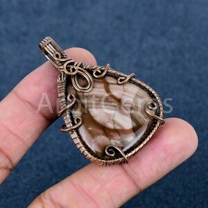 May include: A teardrop-shaped pendant featuring a polished brown and cream-colored stone encased in intricate copper wire wrapping. The metalwork includes decorative coils and a secure bail at the top, with the text ArbitGems visible.