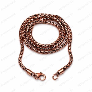 Puede incluir: Un collar de cadena de trigo en tono cobre con un diseño de eslabones trenzados y texturizados. La cadena está enrollada en forma circular e incluye un cierre de mosquetón en un extremo y una pequeña anilla circular en el otro.