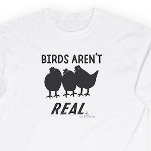 Op de afbeelding: Een wit t-shirt met een zwarte afbeelding van drie vogels op een rij. Boven de vogels staat de tekst "BIRDS AREN'T" in zwarte hoofdletters. Onder de vogels staat de tekst "REAL" in het zwart, gevolgd door "SEE the TRUTH".