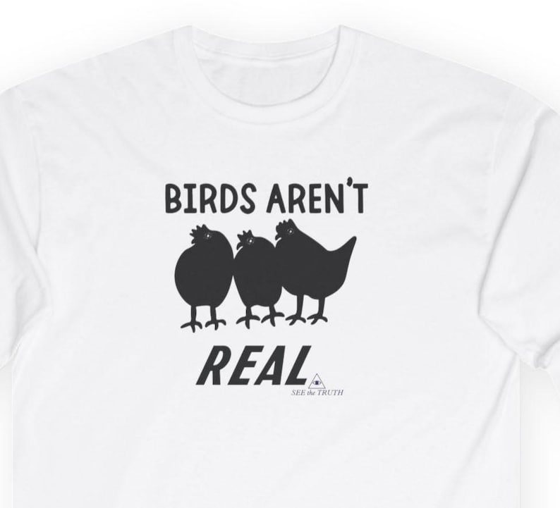 Op de afbeelding: Een wit t-shirt met een zwarte afbeelding van drie vogels op een rij. Boven de vogels staat de tekst "BIRDS AREN'T" in zwarte hoofdletters. Onder de vogels staat de tekst "REAL" in het zwart, gevolgd door "SEE the TRUTH".