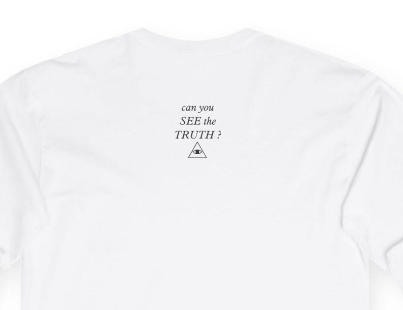 Op de afbeelding: Een effen wit shirt met lange mouwen en zwarte tekst in het midden op de bovenrug die luidt can you SEE the TRUTH? boven een kleine zwarte lijntekening van een driehoek met een oogsymbool. De stof is glad en volledig wit van kleur.