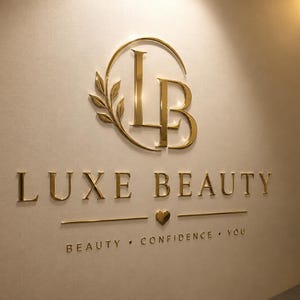 May include: A gold-colored 3D wall sign featuring the letters LB intertwined with a leaf branch inside a circle. Below, the text LUXE BEAUTY appears in gold lettering, followed by a small heart icon and the words BEAUTY &bull; CONFIDENCE &bull; YOU.