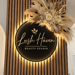 以下が含まれることがあります： Lush Haven BEAUTY STUDIOと金色の文字で書かれた円形の黒い看板。温かみのあるバックライトで照らされています。看板は垂直の木製スラット壁に取り付けられており、乾燥したパンパスグラスと花の大きなアレンジメントで装飾されています。