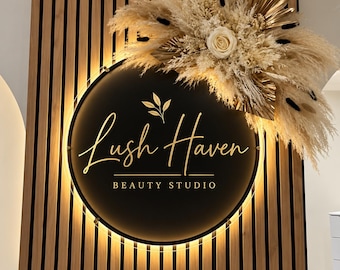 May include: A circular black sign with gold lettering reading Lush Haven BEAUTY STUDIO, illuminated by a warm backlight. The sign is mounted on a vertical wood slat wall and decorated with a large arrangement of dried pampas grass and flowers.