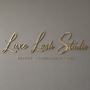 May include: A gold-colored wall sign featuring the text Luxe Lash Studio in a cursive script font, with the words BEAUTY &bull; CONFIDENCE &bull; YOU in a smaller, sans-serif capital letter font positioned directly underneath the main script text.