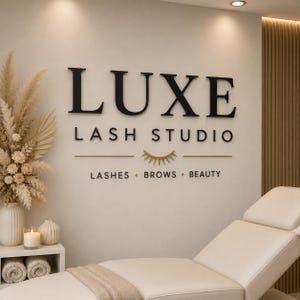 May include: A wall sign reading LUXE LASH STUDIO with a gold eyelash icon and the text LASHES BROWS BEAUTY. The sign is mounted on a light-colored wall above a white treatment bed, with a vase of dried flowers and a candle on a side table.