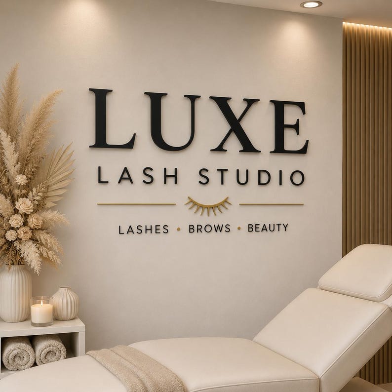 May include: A wall sign reading LUXE LASH STUDIO with a gold eyelash icon and the text LASHES BROWS BEAUTY. The sign is mounted on a light-colored wall above a white treatment bed, with a vase of dried flowers and a candle on a side table.