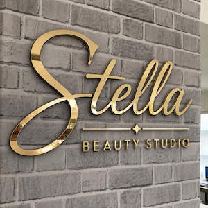 May include: A three-dimensional wall sign featuring the word Stella in a large, gold, reflective script font, positioned above the text BEAUTY STUDIO in a smaller, gold, sans-serif font, separated by a horizontal line with a central star.