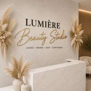 May include: A wall sign for Lumi&egrave;re Beauty Studio featuring black and gold 3D lettering. The text reads Lumi&egrave;re Beauty Studio and LASHES &bull; BROWS &bull; SKIN &bull; CONFIDENCE. The wall is decorated with two large arrangements of dried pampas grass and palm leaves.