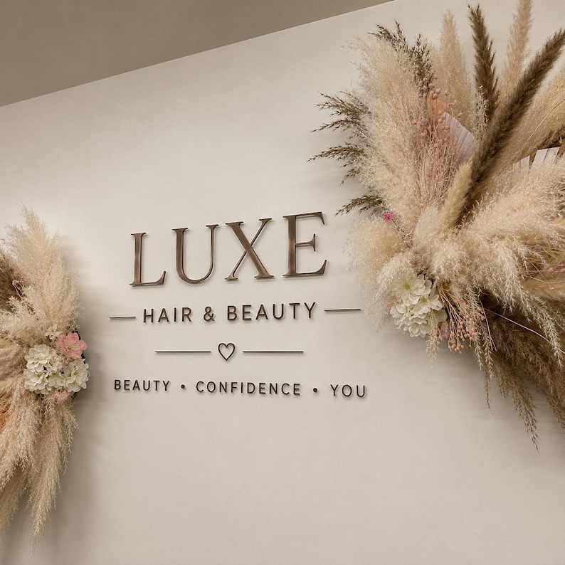 May include: A wall sign featuring the text LUXE HAIR & BEAUTY, BEAUTY &bull; CONFIDENCE &bull; YOU, and a small heart symbol. The sign is flanked by two large, decorative arrangements of dried pampas grass, beige plumes, and white floral accents.