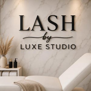 May include: A wall sign with black 3D lettering reading LASH by LUXE STUDIO mounted on a white marble wall. Below the sign is a white treatment bed with a beige blanket, and a small white shelf holding a vase with dried grass and black bottles.