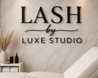 May include: A wall sign with black 3D lettering reading LASH by LUXE STUDIO mounted on a white marble wall. Below the sign is a white treatment bed with a beige blanket, and a small white shelf holding a vase with dried grass and black bottles.
