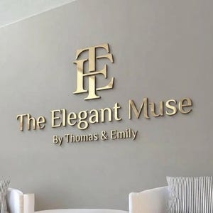 May include: A three-dimensional wall sign featuring a gold-coloured metallic monogram of the letters T and E, followed by the text The Elegant Muse and the smaller text By Thomas & Emily, all mounted on a smooth, light grey wall surface.