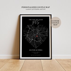 Puede incluir: Un cuadro decorativo con marco negro que muestra un mapa de calles blanco con forma de corazón. El texto dice THE DAY WE SAID YES, OLIVER & EMMA y THE PLACE OUR STORY BEGAN, con un pequeño icono de corazón rojo marcando una ubicación.