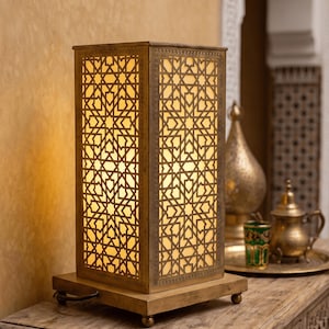 Op de afbeelding: Een rechthoekige tafellamp met een goudkleurig metalen frame met ingewikkelde geometrische uitsparingen. De lamp verspreidt een warme, zachte gloed van binnenuit, waardoor de gedetailleerde patronen worden verlicht. Het rust op een vierkante voet met vier kleine pootjes.