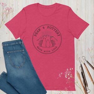 Puede incluir: Una camiseta roja jaspeada con un logotipo en blanco y negro que dice "Peak 6 Pottery Made With Love". El logotipo presenta una cordillera con un sol saliendo detrás y tres jarrones de cerámica.