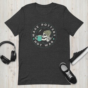 Könnte beinhalten: Dunkelgraues T-Shirt mit einem Totenkopf, der eine Mütze trägt und aus einer Tasse trinkt. Der Totenkopf hält ein Peace-Zeichen hoch. Der Text auf dem T-Shirt lautet "Make Pottery Not War".