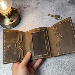 May include: A tri-fold brown leather wallet featuring a distressed texture, white contrast stitching, multiple card slots, and a central clear ID window. The wallet includes a silver metal snap button closure and a visible interior layout.