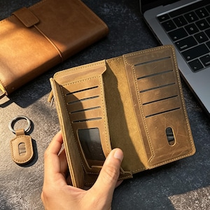 May include: Open, tan leather wallet with several card slots and a transparent ID window. A matching leather keyring and a closed wallet are also visible. The wallet is held open, displaying its internal design.