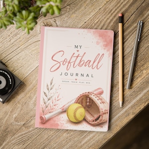 May include: A rectangular journal with a light pink cover featuring the text MY Softball JOURNAL and DREAM. TRAIN. PLAY. WIN. The cover displays a watercolour illustration of a softball, a bat, a glove, and leafy branches in shades of pink.