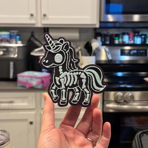 May include: A black embroidered patch featuring a stylized unicorn with a white skeletal rib cage, spine, and skull design. The unicorn has a prominent horn, a flowing mane, and a tail, all outlined in white thread against a dark fabric background.