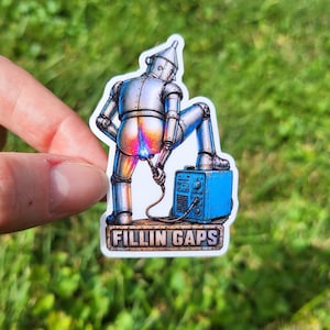 May include: A die-cut sticker featuring a silver tin-man character welding its own metallic backside with a blue welding machine. The character stands on a platform with the text FILLIN GAPS printed in bold, blocky, weathered-style lettering.